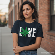 Woman wearing a black t-shirt with a cannabis leaf design and 'LOVE' text, standing in front of a brick building.