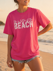 Woman wearing a pink 'Life is better at the beach' t-shirt on a beach.