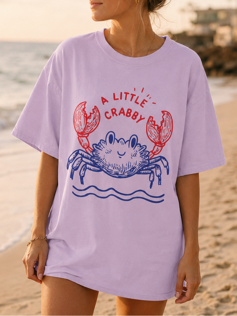 Woman wearing a light purple t-shirt with a crab graphic on a beach at sunset.