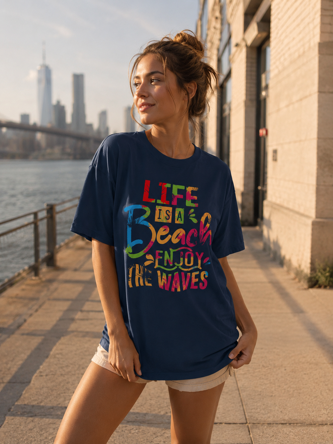 Woman wearing a navy blue t-shirt with colorful text, standing by a waterfront with a city skyline in the background.