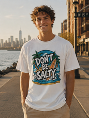 Man wearing a white t-shirt with 'Don't Be Salty' graphic design by a waterfront with city skyline.