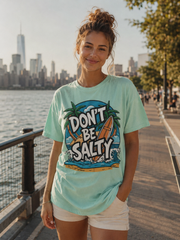 Woman wearing a 'Don't Be Salty' t-shirt by a waterfront with a city skyline in the background.
