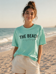 Woman wearing a light blue t-shirt with 'The Beach is my Therapy' text on a beach.