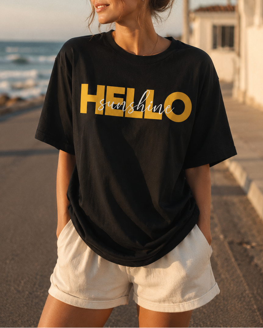 Woman wearing a black t-shirt with 'HELLO sunshine' printed in yellow at the beach.