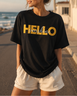 Woman wearing a black t-shirt with 'HELLO sunshine' printed in yellow at the beach. thumbnail 1