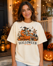 Female model wearing the Comfort Colors 1717 Ivory Halloween Is Good skeleton pumpkin chair T-shirt