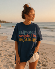Woman wearing a 'saltwater seashells sunshine' t-shirt on a beach.